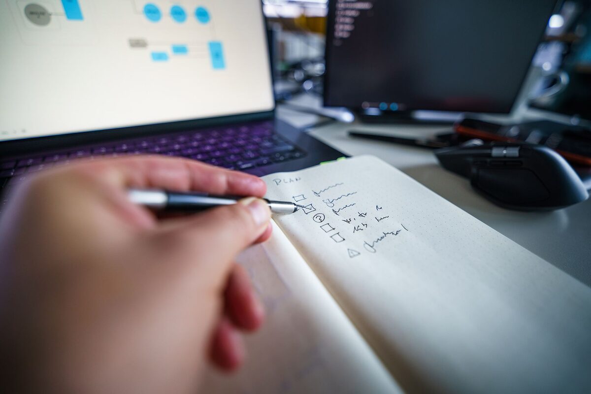 Dłoń notująca w zeszycie obok otwartego laptopa